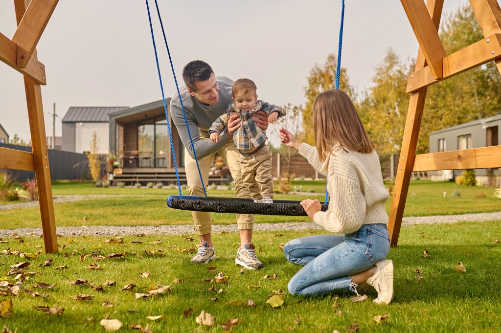 качели family-leisure-young-adult-smiling-man-supporting-child-swing-woman-crouching-with-outstretched-hand-spending-leisure-time-together-near-country-house-autumn-day.jpg
